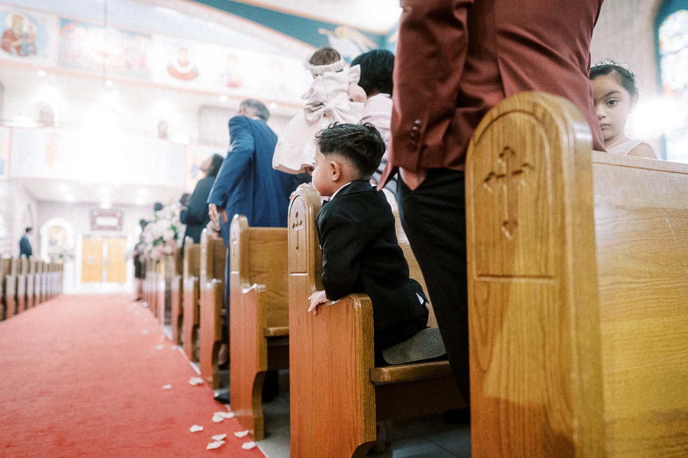 A young boy in a suit stands on a church pew, surrounded by elegantly dressed people, during a wedding ceremony. The church
