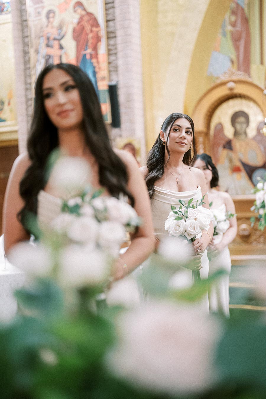 Bridesmaid holding a bouquet of white roses stands in a church setting adorned with religious icons, capturing a serene and