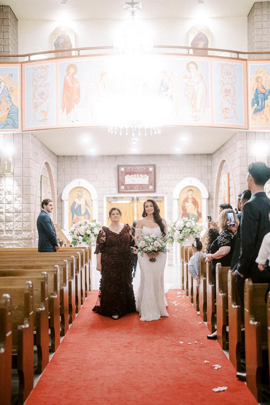 A bride in a white wedding gown walks down the aisle with a woman in burgundy, surrounded by guests in a church with