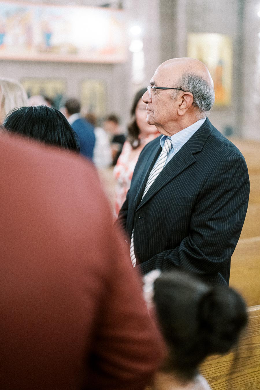 Older gentleman in a suit attending a formal event inside a church, surrounded by other attendees in a softly lit setting.