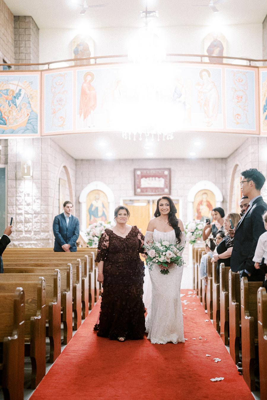 Bride in elegant white gown walks down the red carpet aisle of a beautifully decorated church, accompanied by a woman in a