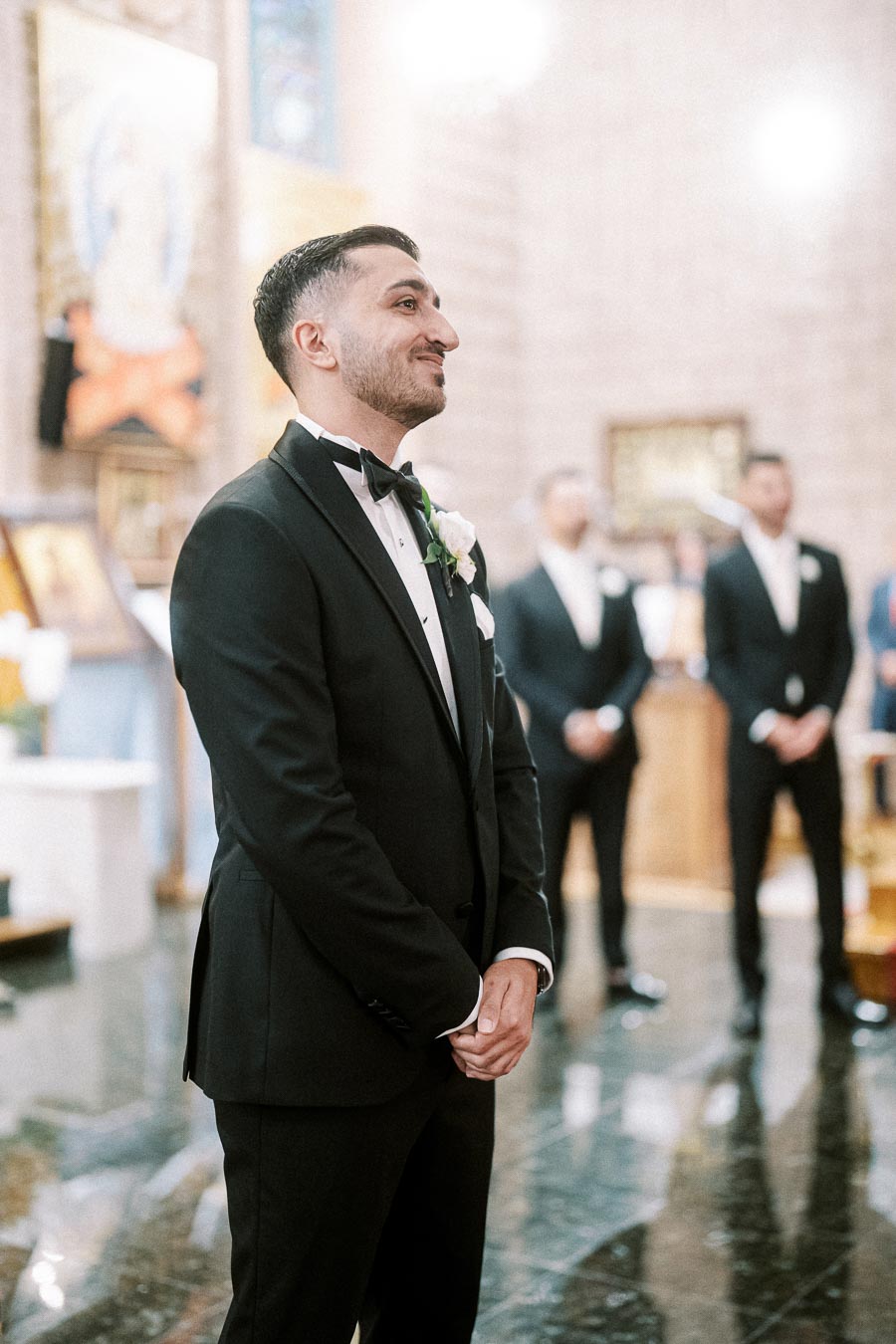 Groom standing in a church, wearing a black tuxedo and bow tie, with groomsmen in the background, smiling during a wedding