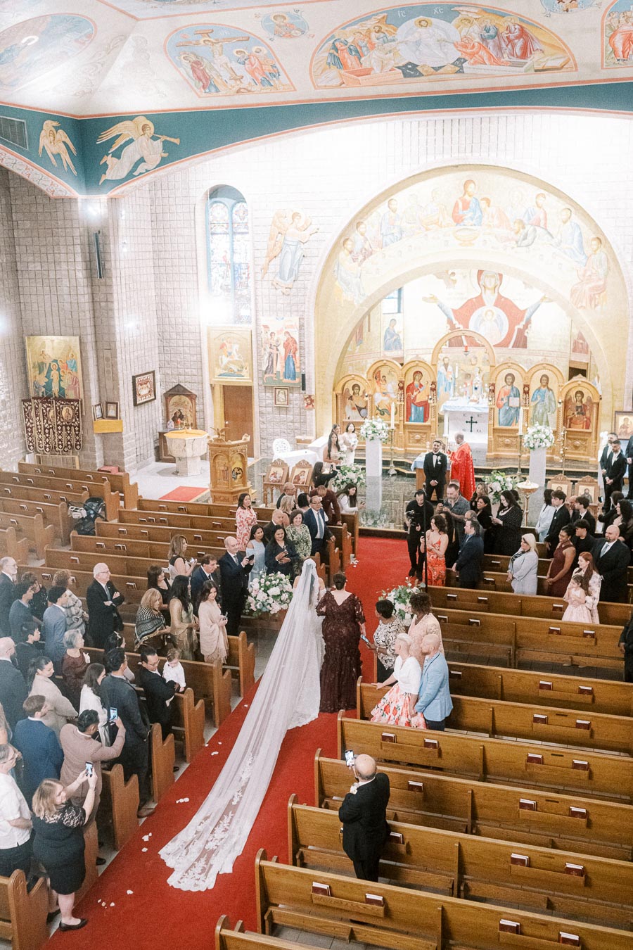 Aerial view of a wedding ceremony inside an ornately decorated church, with a bride in a long, flowing veil walking down the