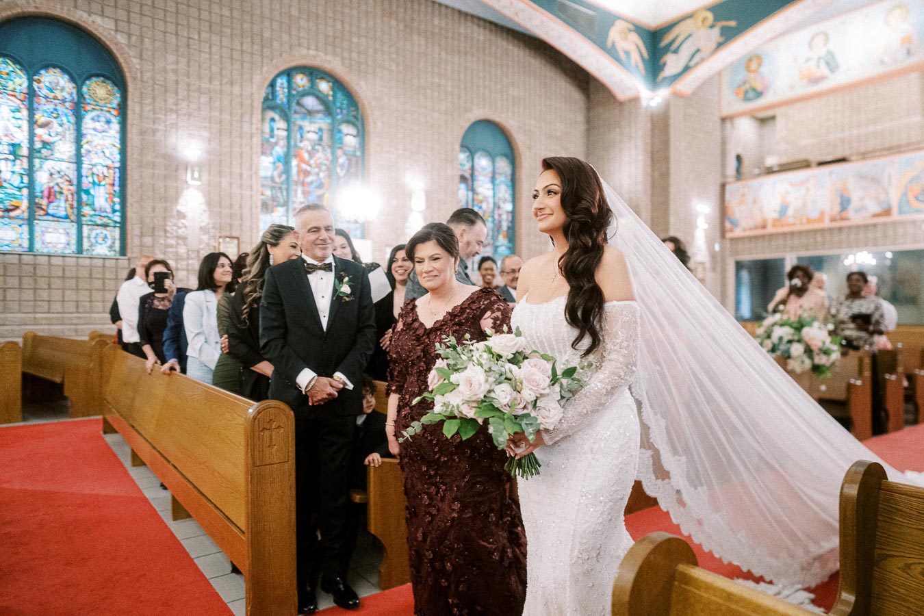 Bride walking down the aisle with parents in a beautifully decorated church, surrounded by guests and adorned with stained