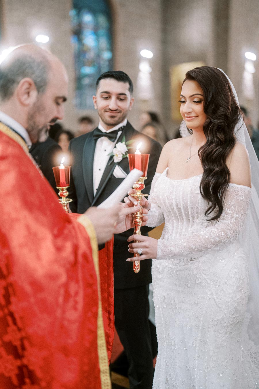 Elegant wedding ceremony in a church, featuring a bride in a lace gown and a groom in a tuxedo holding red candles, with a