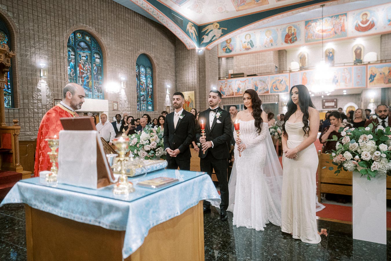 A wedding ceremony in a beautifully decorated church with stained glass windows, featuring a couple holding candles and
