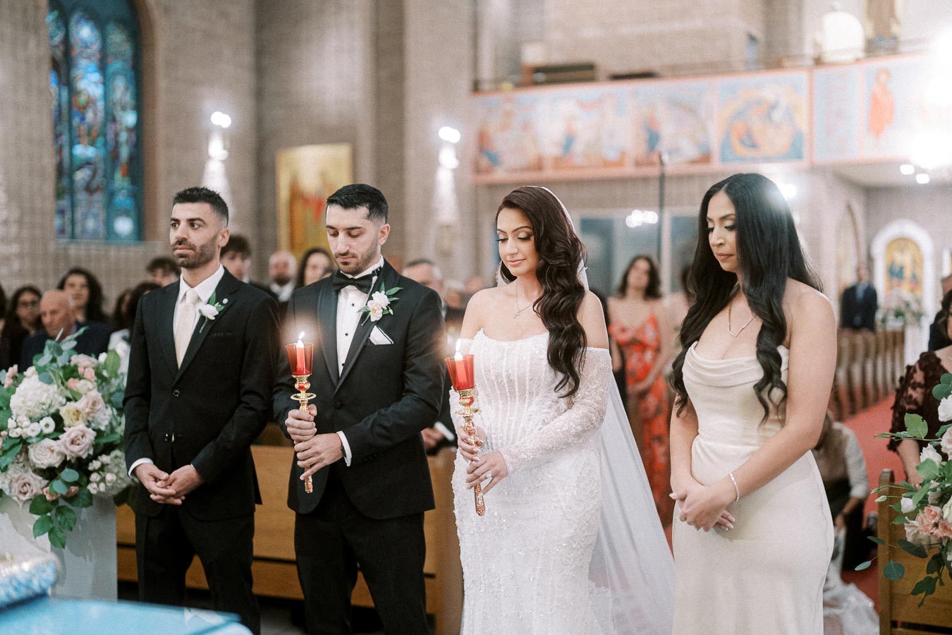 Wedding ceremony in a beautifully decorated church with bride and groom holding red candles, accompanied by bridesmaid and