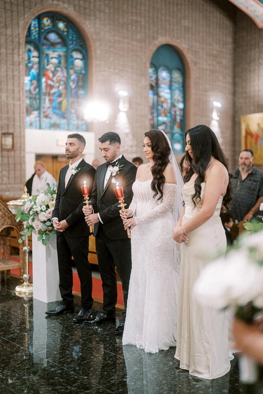 A wedding ceremony inside a beautifully decorated church with stained glass windows; a bride in an elegant white gown stands