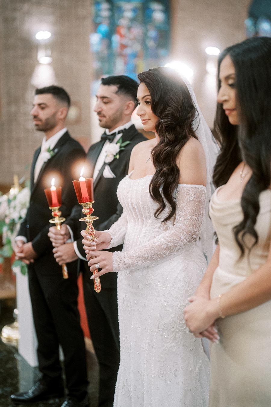 A bride and groom holding candles during a traditional wedding ceremony, surrounded by groomsmen and bridesmaids, in an