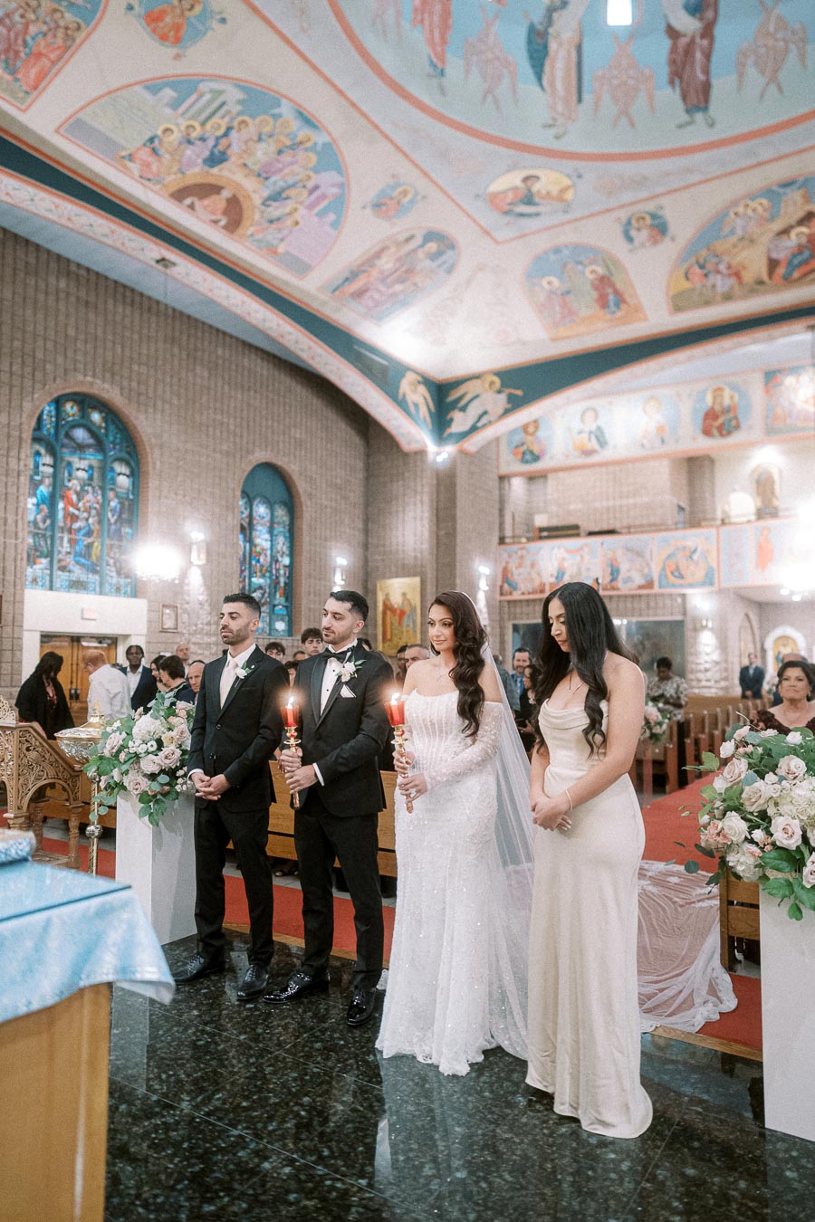A couple holding candles stands in a beautifully decorated church during a wedding ceremony, surrounded by bridesmaids in