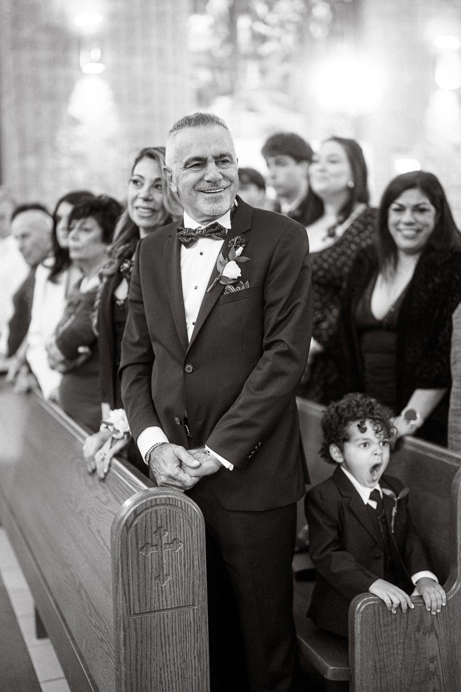 A joyful man in a suit stands in a church aisle, surrounded by smiling family members, as a young boy in a suit yawns beside