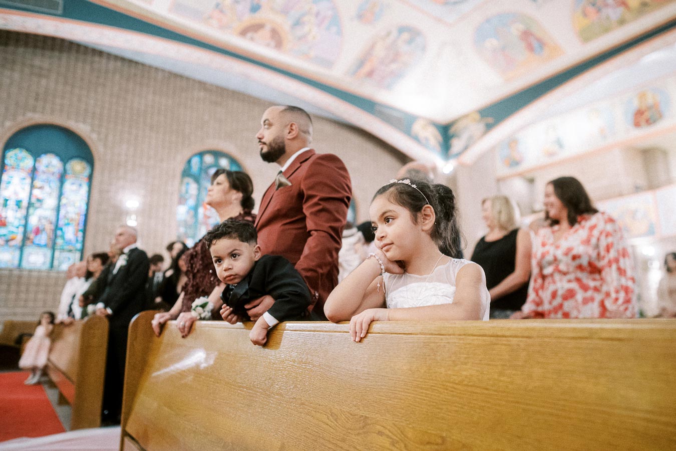 A child in a white dress looks thoughtful while leaning on a church pew, surrounded by family members attending a ceremony