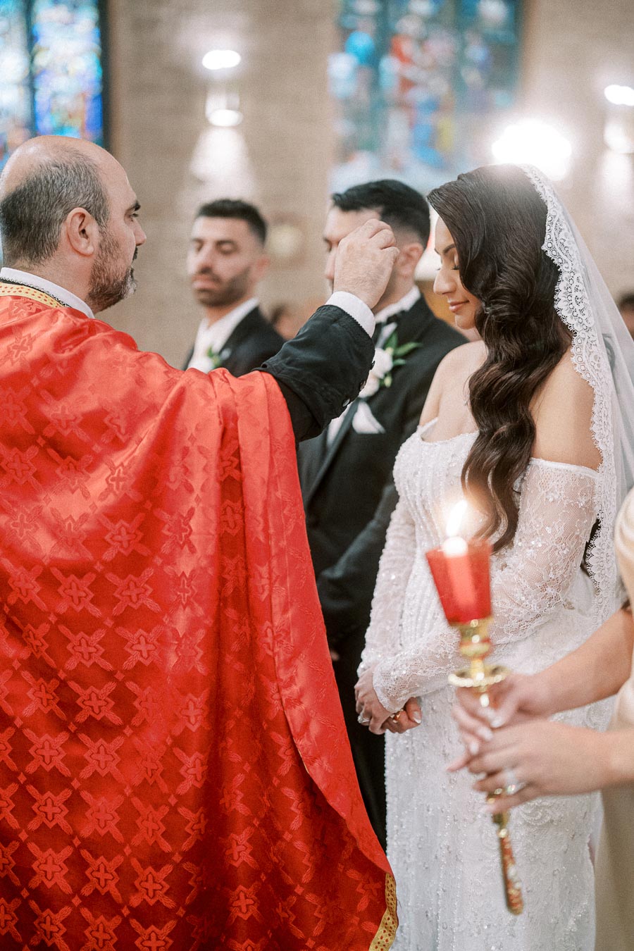 A bride and groom receiving a blessing from a priest during a traditional wedding ceremony in a church with stained glass