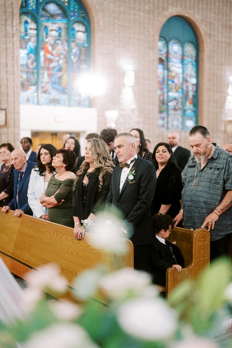 A group of elegantly dressed people standing in a church during a wedding ceremony, with stained glass windows in the