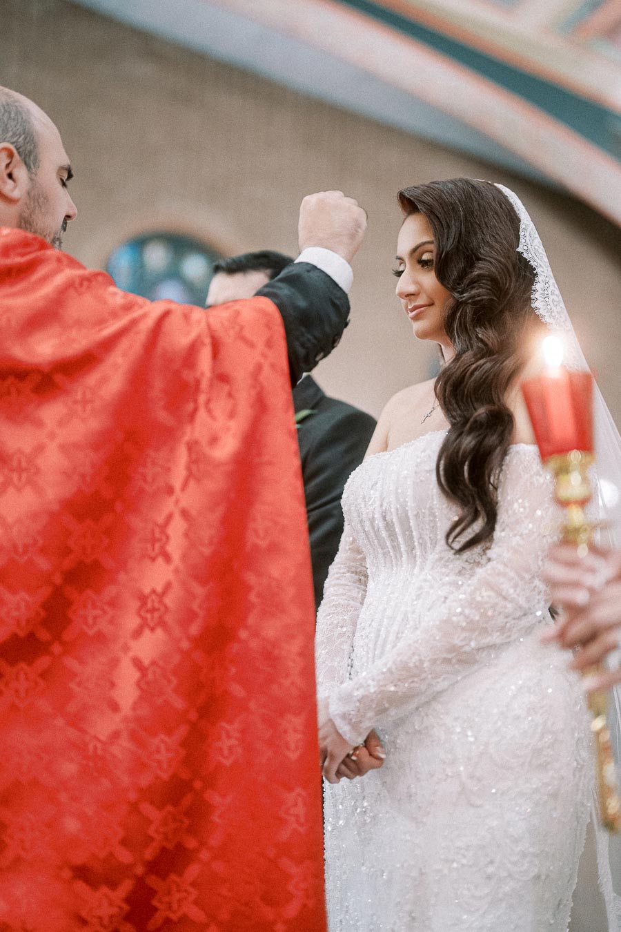 A bride in an elegant white lace gown participating in a traditional wedding ceremony, standing beside a person in a red