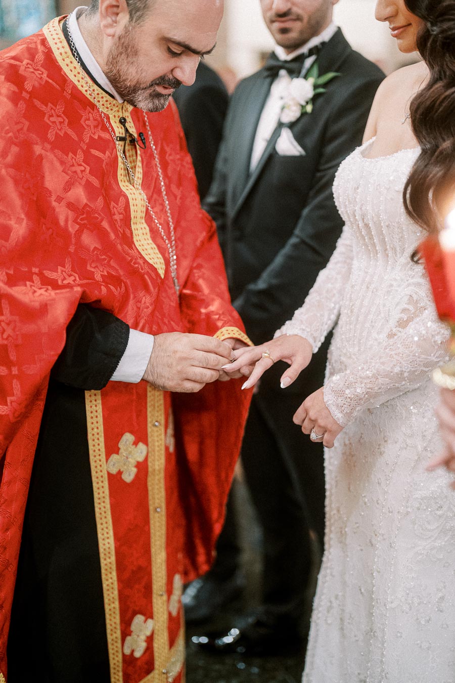 A priest in a red robe officiates a wedding ceremony, placing a ring on the bride's finger, with the groom standing nearby