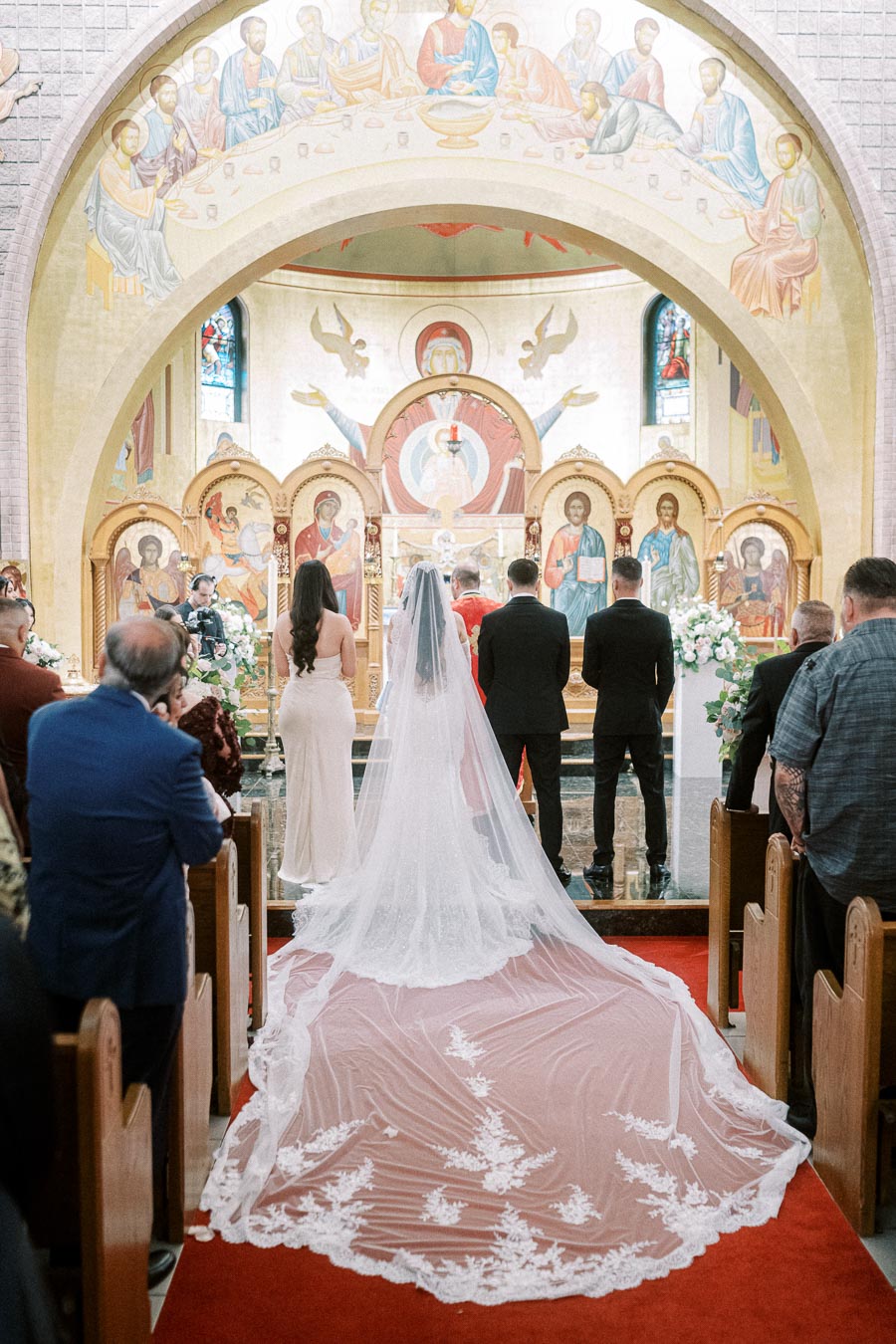 A bride in a long, elegant veil stands at the altar with the groom in a beautifully decorated church, watched by guests
