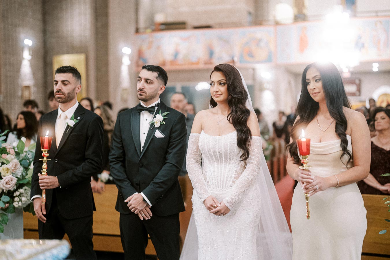 A bride and groom stand during their wedding ceremony in a beautifully decorated church, accompanied by two attendants