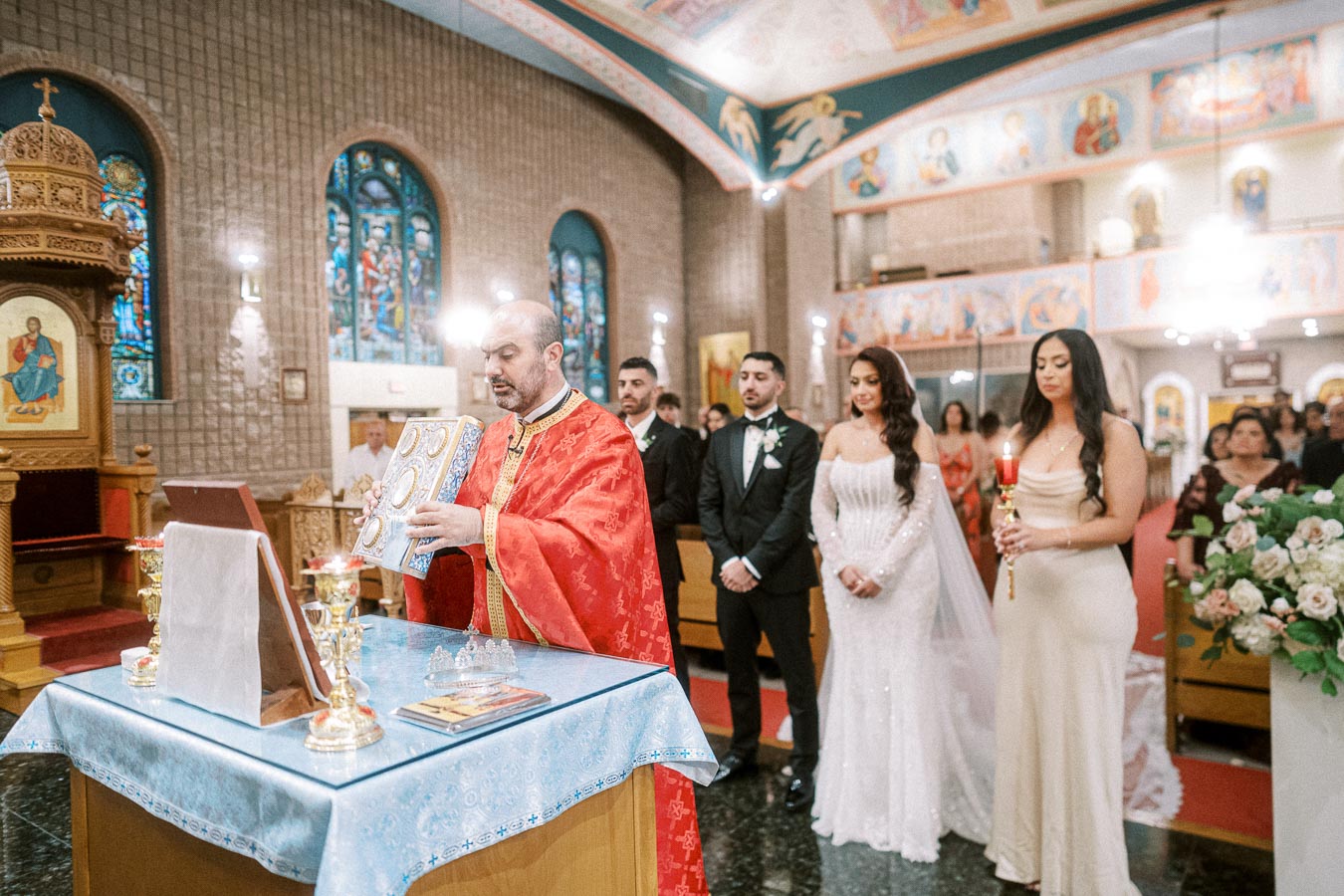 Orthodox wedding ceremony inside a beautifully adorned church, featuring a priest in red robes holding a religious book,