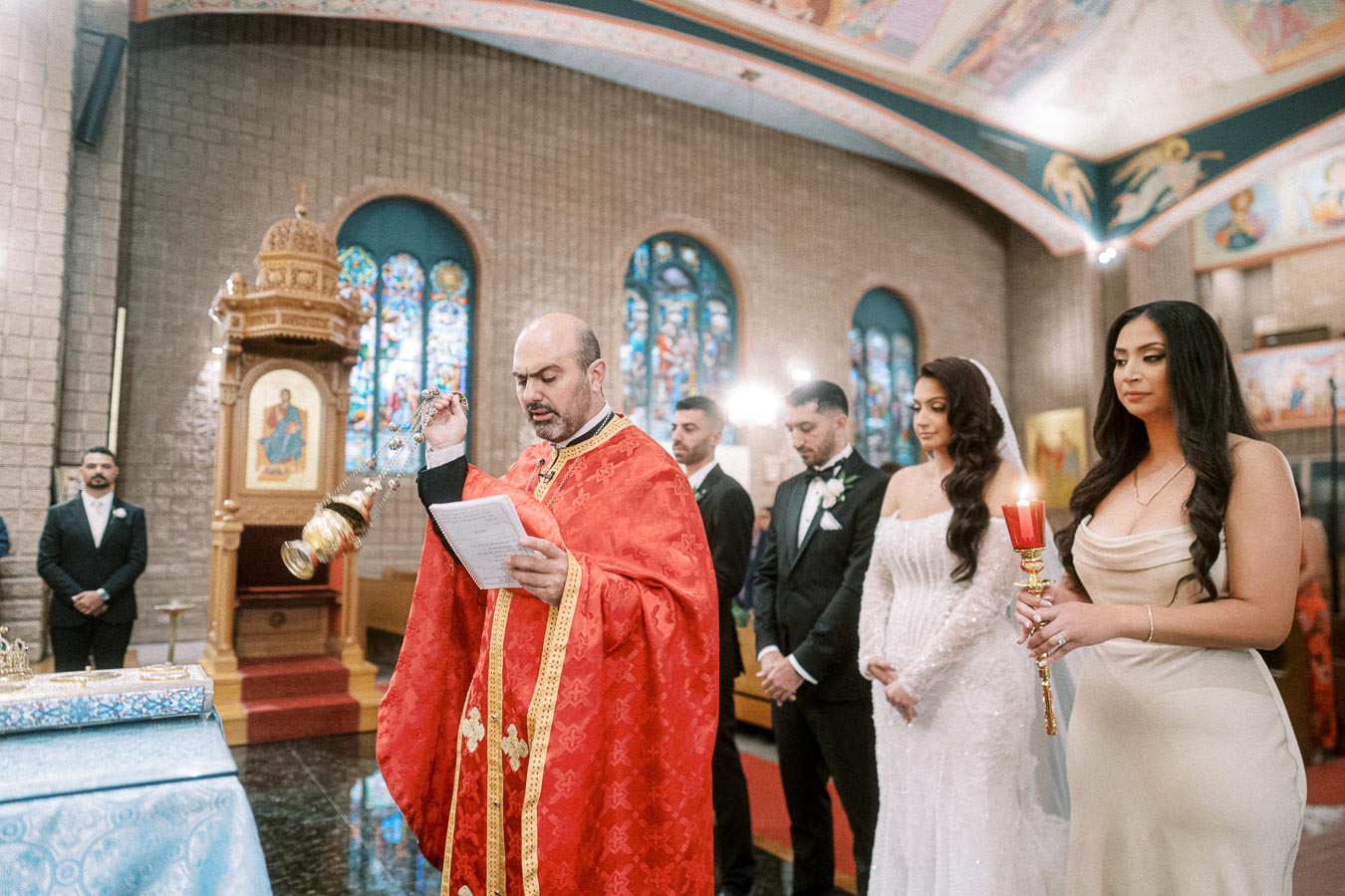 A priest in red robes conducting a traditional wedding ceremony in a church with ornate stained glass windows, while the