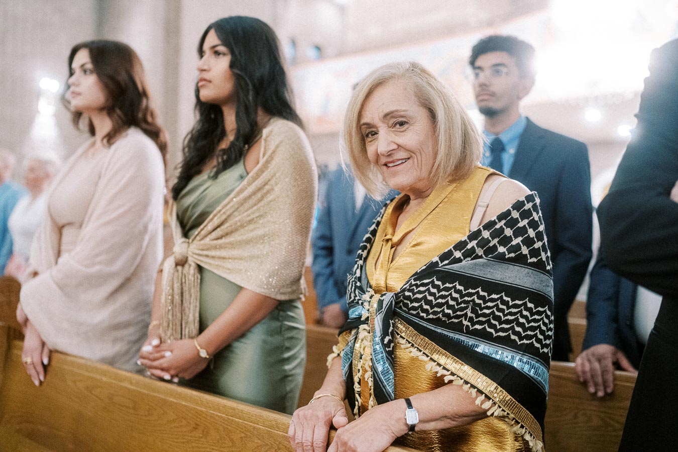 A group of people dressed elegantly standing in a church pew, with one older woman smiling and wearing a colorful shawl at a