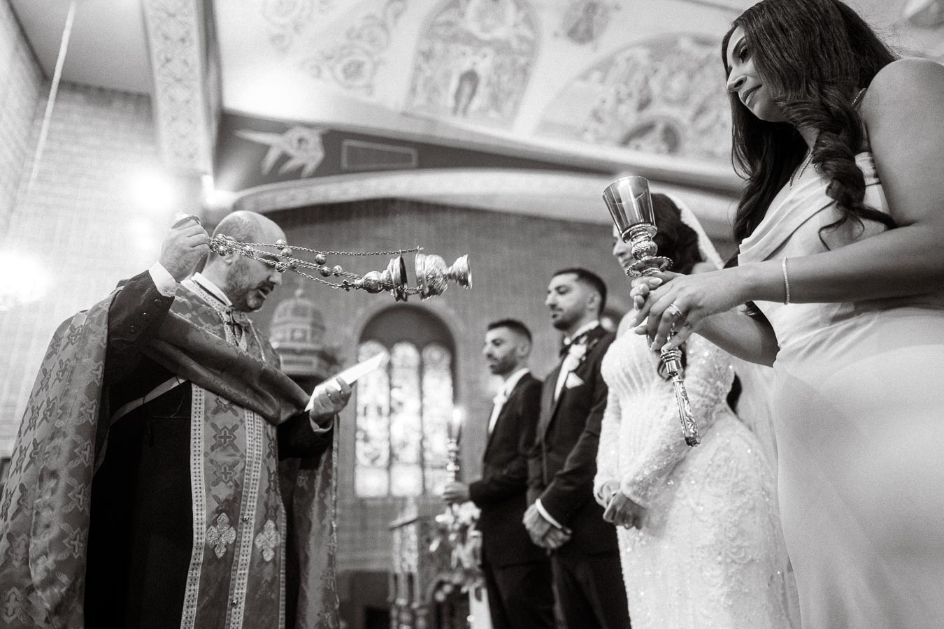 Black and white image of a traditional wedding ceremony in a church, featuring a priest with incense and a row of