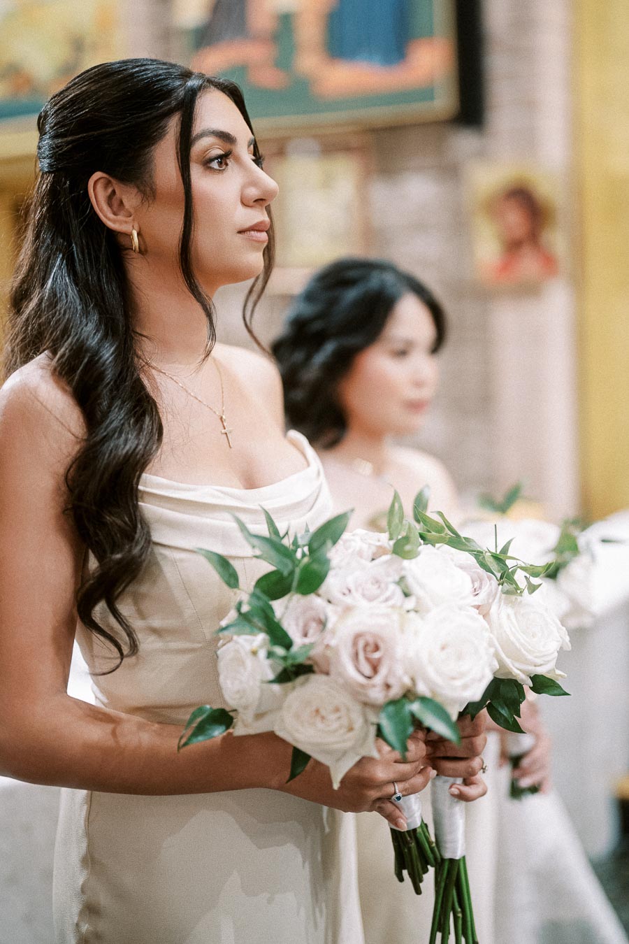 A bridesmaid in an elegant cream dress holds a bouquet of white and blush pink roses during a wedding ceremony in a church