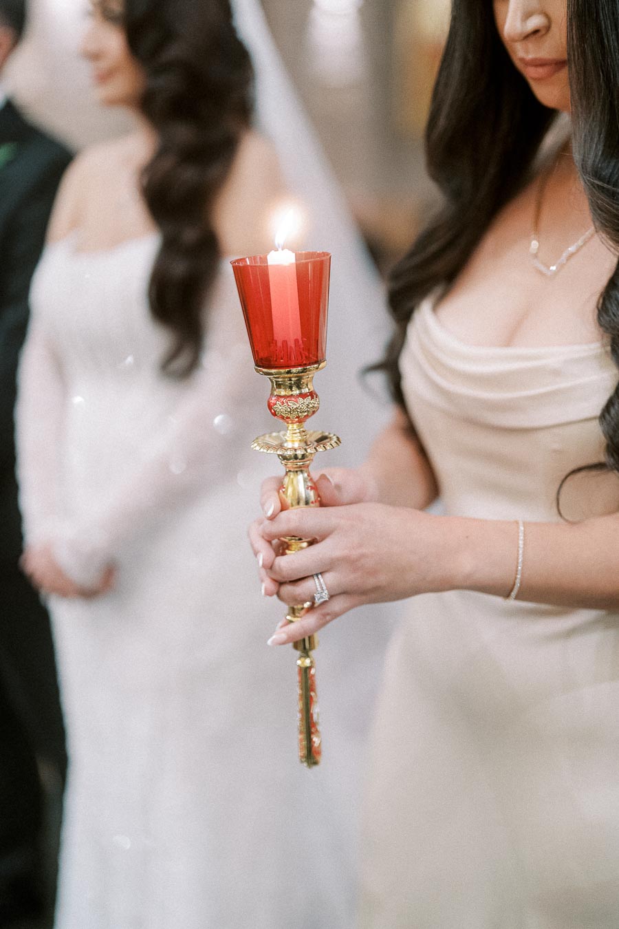 Bridesmaid holding lit red candle during elegant wedding ceremony with bride in background.