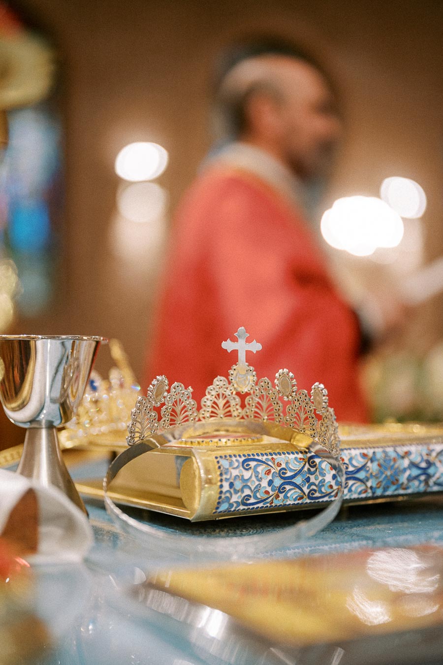 Golden wedding crown with intricate designs placed on ornate book, with priest in red robe blurred in the background,