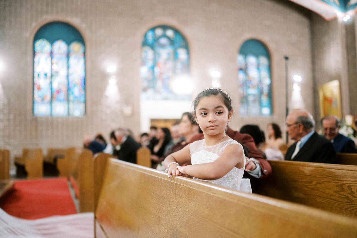 A young girl in an elegant white dress leans on a wooden pew in a beautifully lit church with colorful stained glass windows