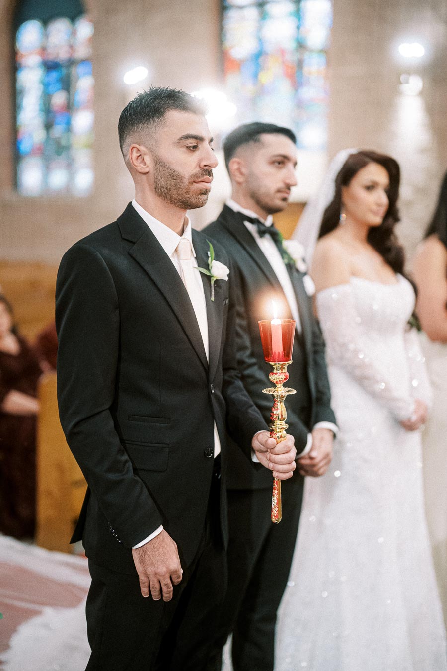 A groom and groomsmen in black tuxedos at a wedding ceremony, with the groom holding a candle, and a bride in a white dress