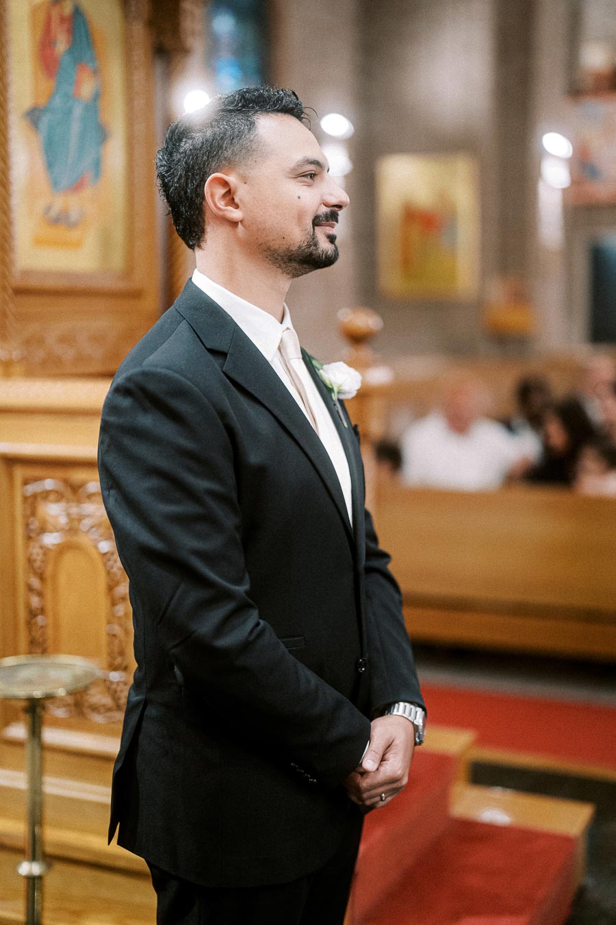 Groom in a tuxedo smiling while standing in a warmly lit church, with religious icons on the wooden altar in the background.