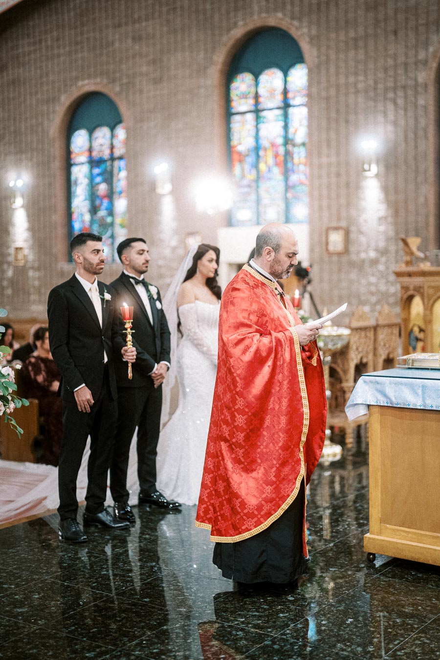 A priest in red ceremonial robes conducts a wedding ceremony in a church with stained glass windows. The bride, groom, and