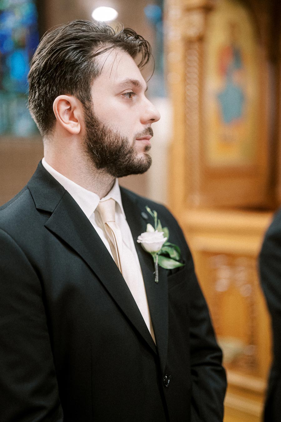 Man in a suit with boutonniere standing indoors, looking attentively to the side, elegant setting in the background