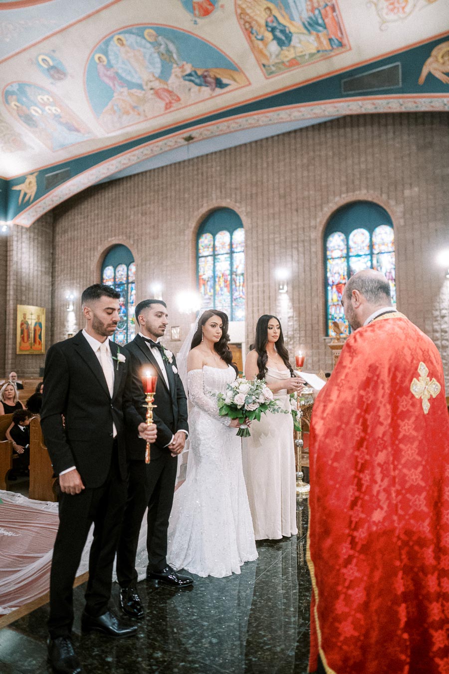 Wedding ceremony in an ornate church with stained glass windows, featuring a bride in a white dress holding a bouquet, groom