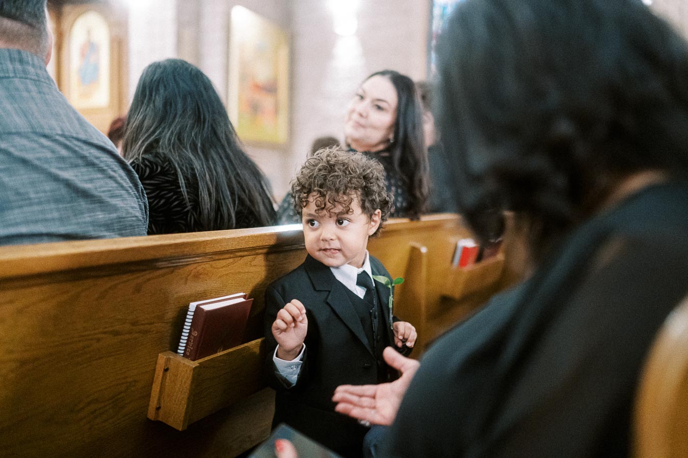 Little boy in a suit sitting in a church pew, looking mischievously backward amidst a wedding ceremony audience.