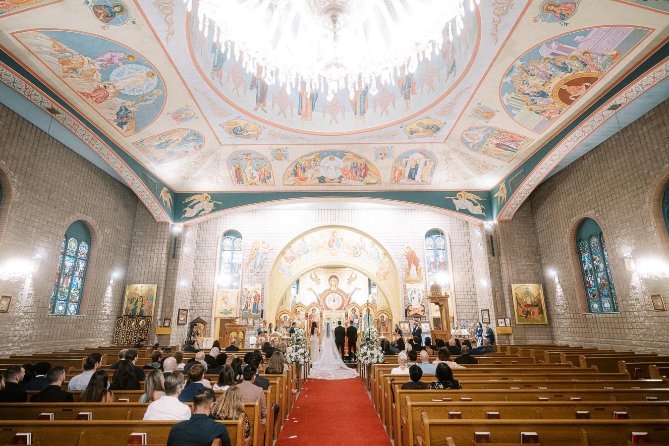 Interior of an ornate church with colorful iconography on the ceiling, a chandelier, stained glass windows, and wedding