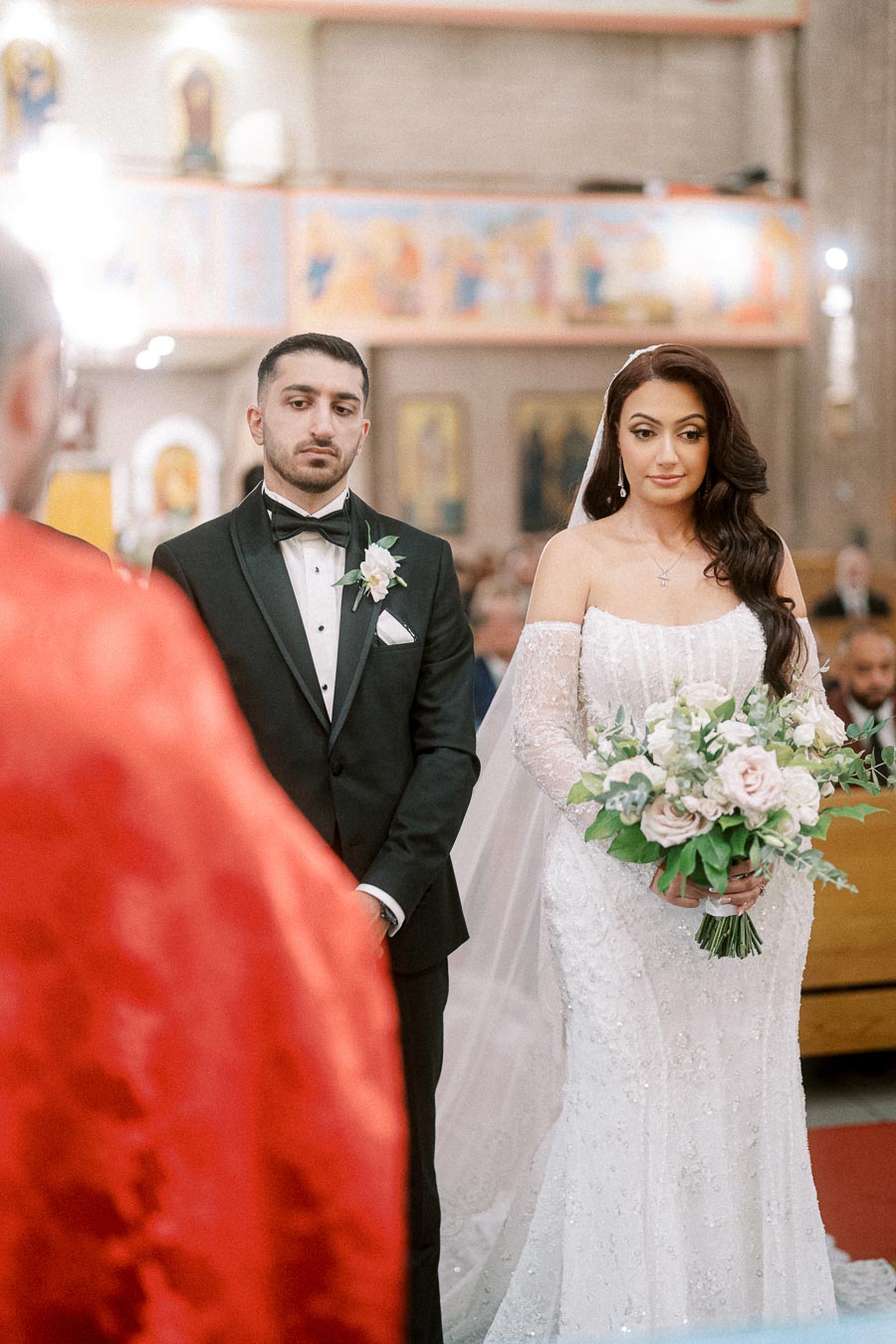 A bride and groom standing during their wedding ceremony in a church, with the bride holding a bouquet of pink and white