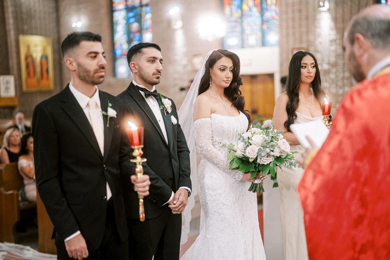 A bride and groom stand at the altar during a wedding ceremony in a church, surrounded by two attendants holding candles.