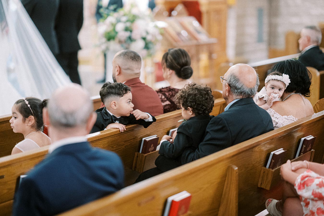A group of people, including children and adults, seated in church pews during a formal event, possibly a wedding, with soft