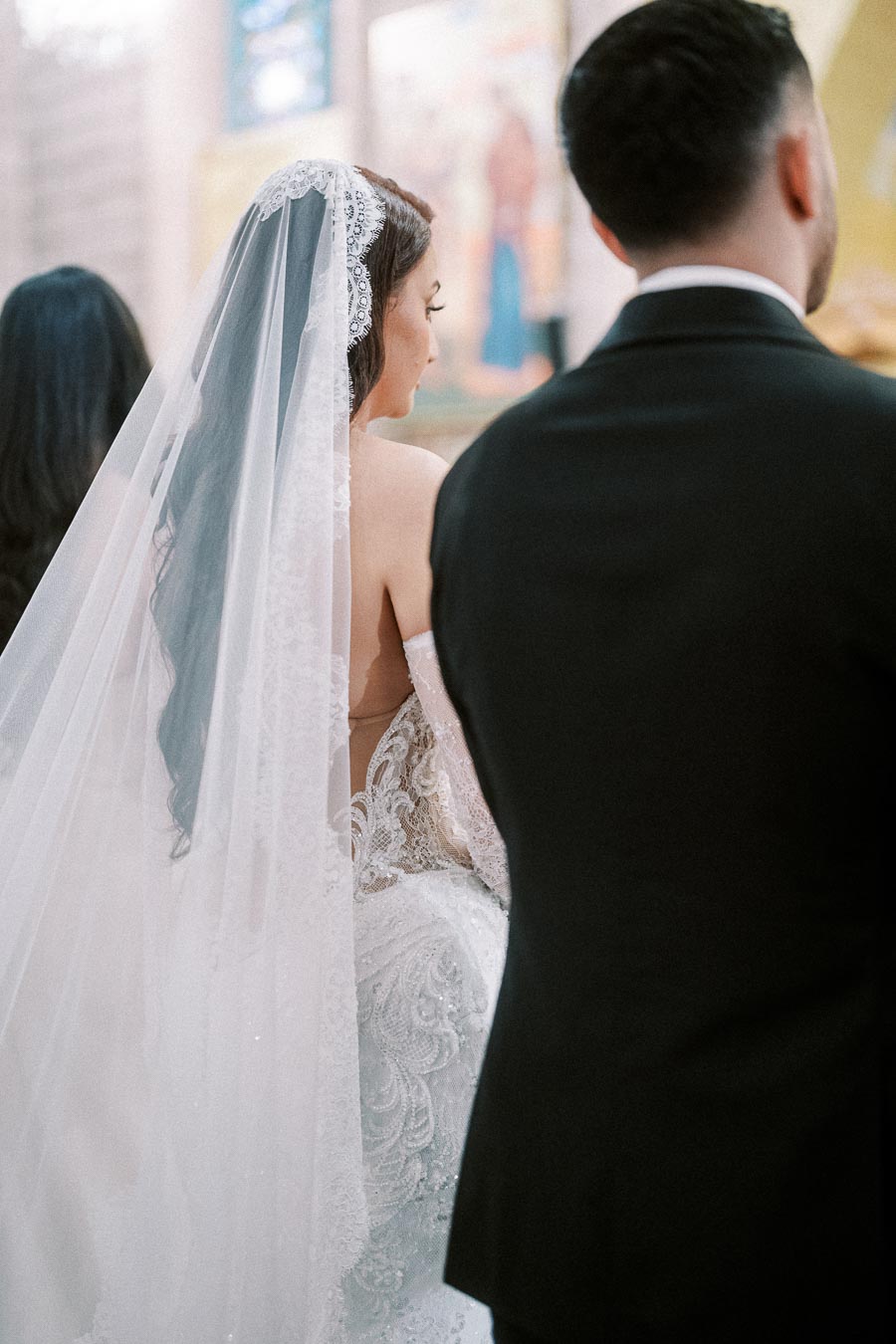 A bride in a lace wedding dress and veil stands beside a groom in a black suit during a wedding ceremony.