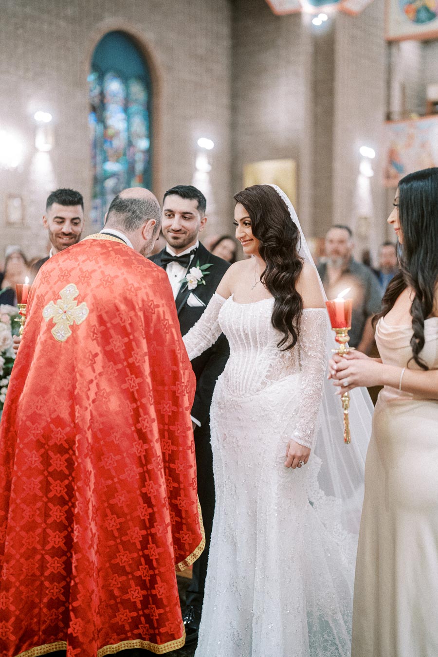 A bride in a white wedding gown and a groom in a black tuxedo stand in a decorated church ceremony. The priest in a red