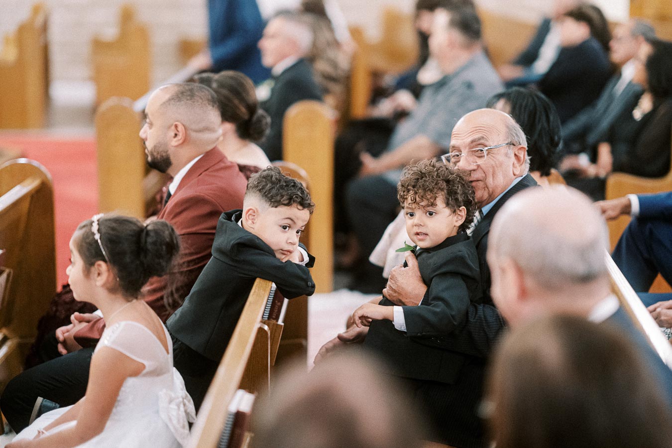 A group of people sitting in a church pew during a formal event, including young children in suits and a girl in a white