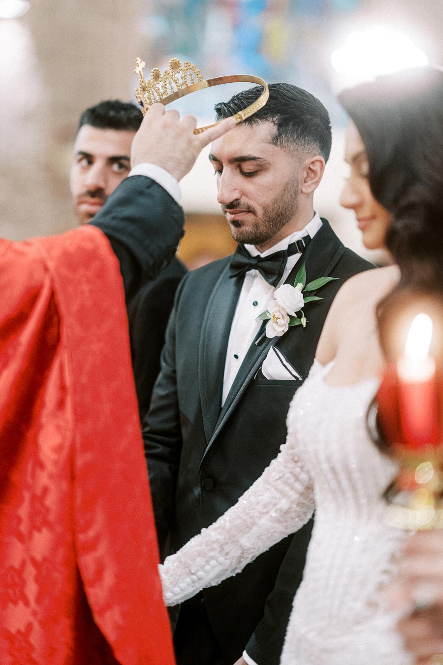 A bride and groom participating in a traditional wedding ceremony, with a priest placing a crown on the groom's head,
