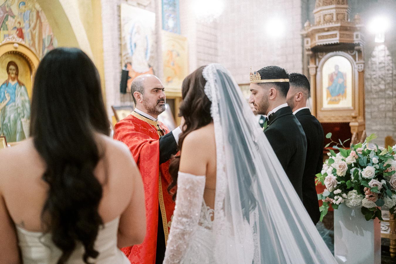 A bride and groom stand before a priest during a traditional wedding ceremony inside a church, surrounded by ornate
