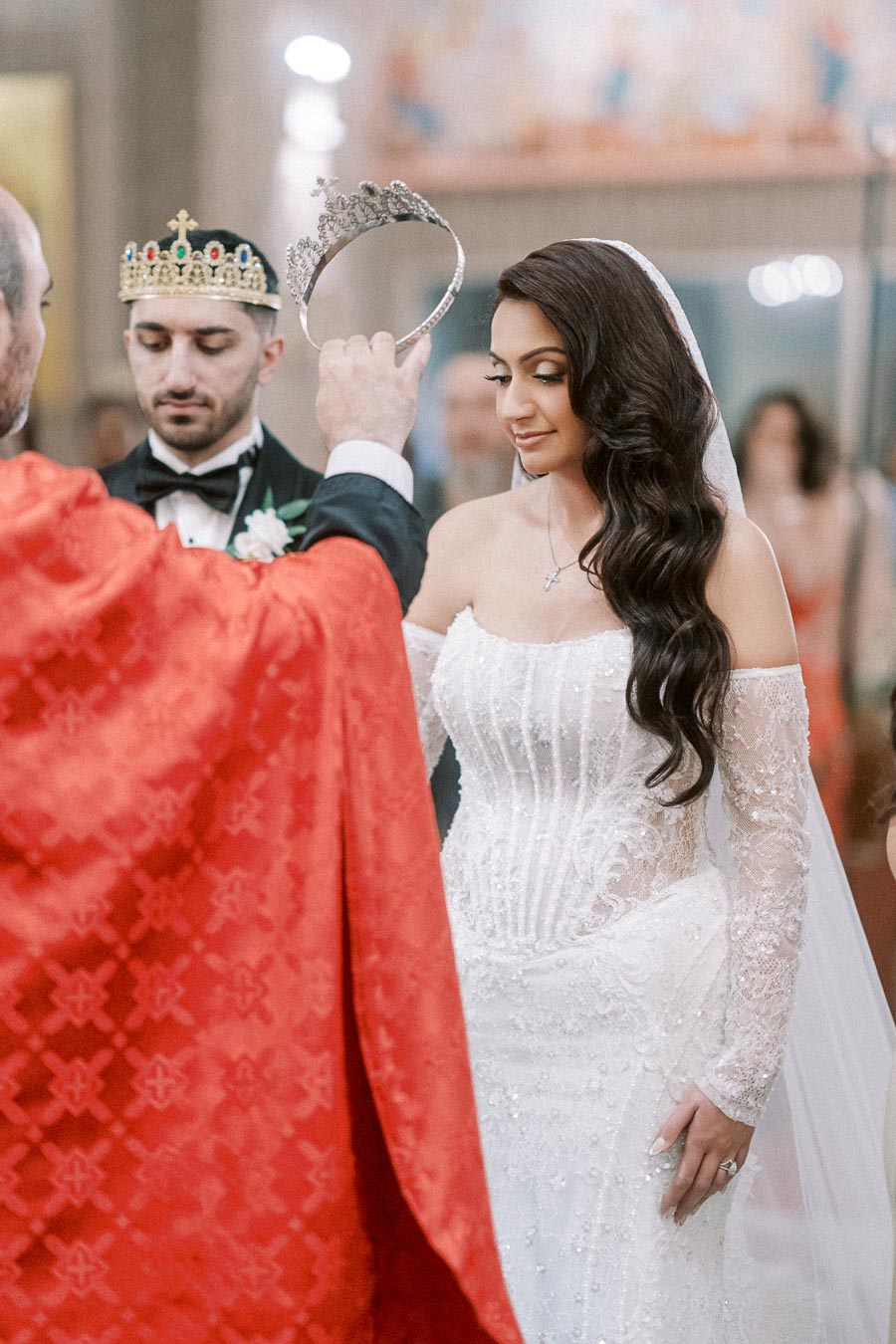 Bride and groom during a wedding ceremony with a priest holding a crown, symbolizing a traditional matrimonial ritual.