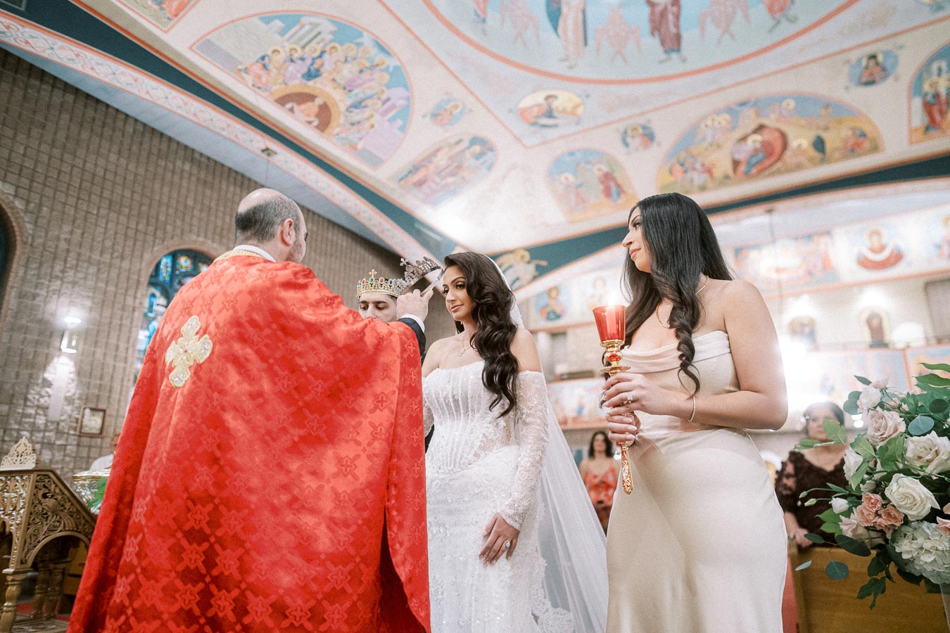 A priest in a red robe conducts a wedding ceremony inside a beautifully decorated church, with the bride in a white gown and