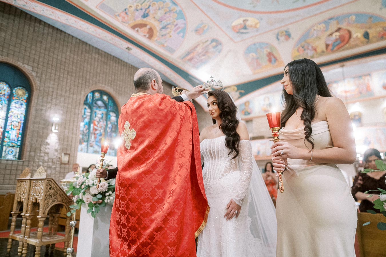A priest in a red robe crowns a bride during a wedding ceremony in a beautifully decorated church, with stained glass