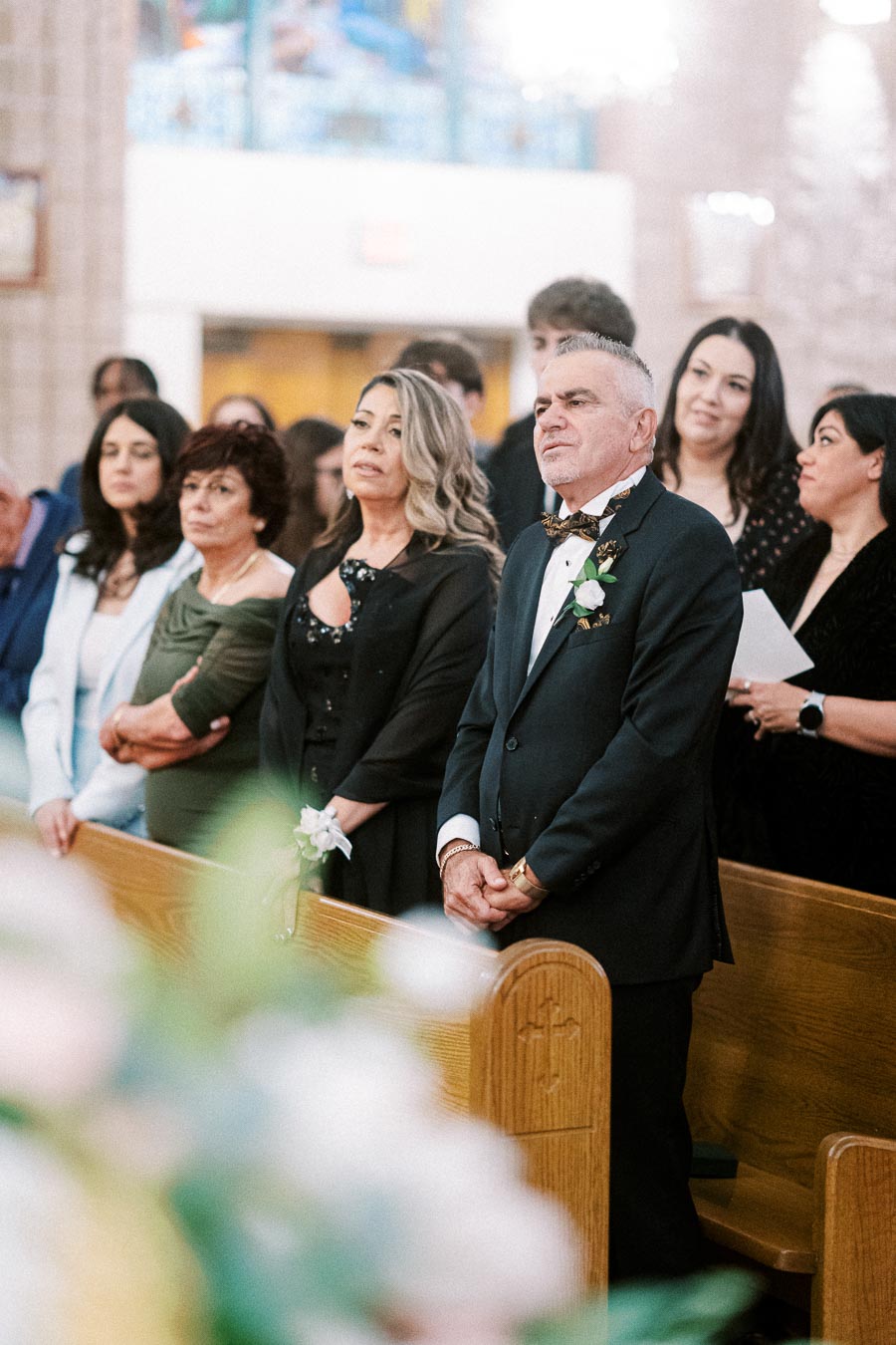 A group of well-dressed people standing in a church during a wedding ceremony, with a man in a suit and bow tie standing