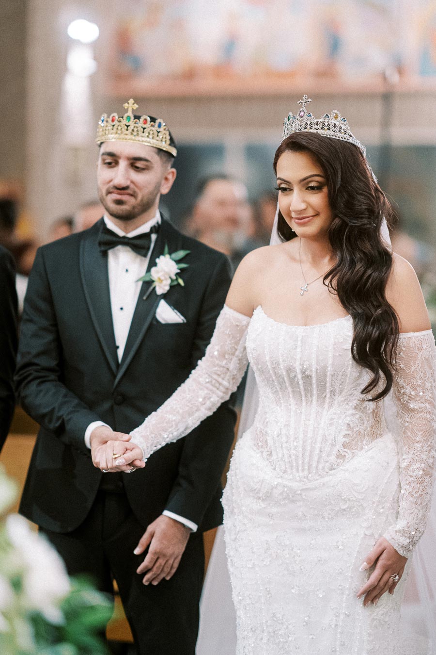 A bride and groom hold hands during their wedding ceremony, both wearing ornate crowns. The bride is in an elegant lace